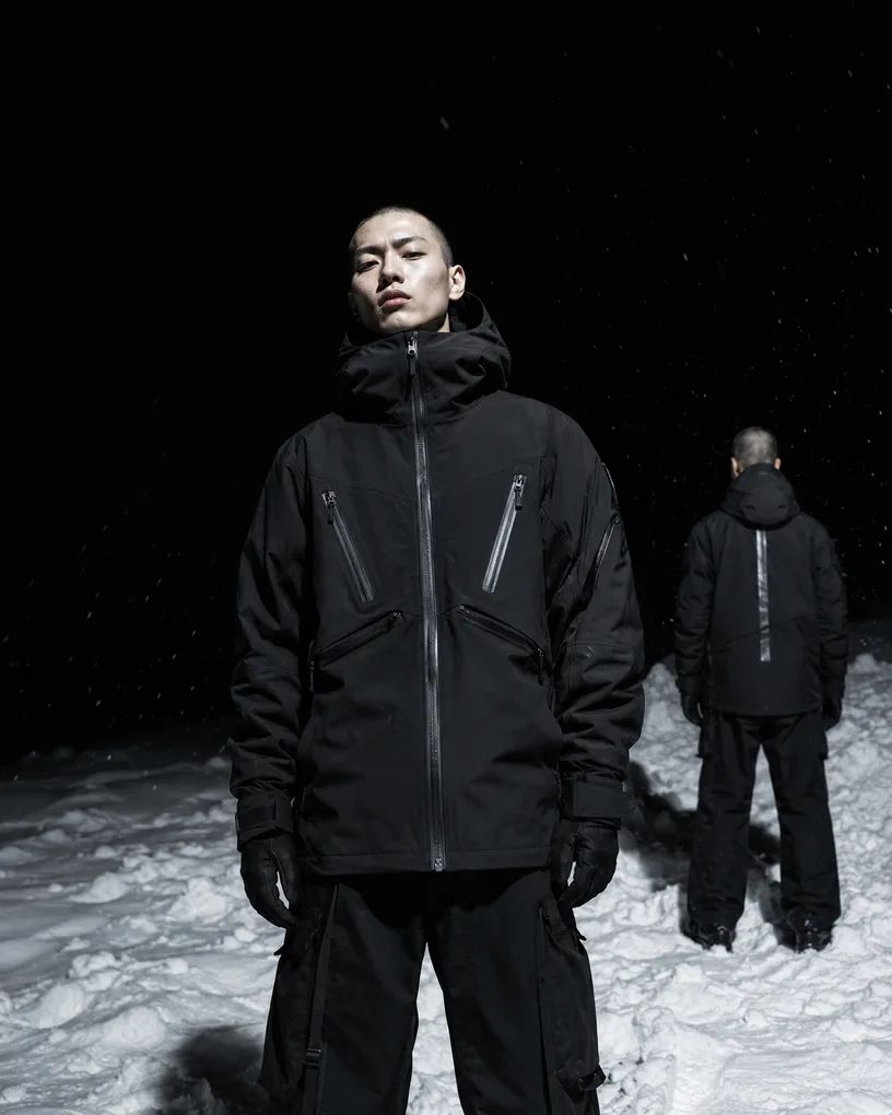 Two figures in black technical jackets in falling snow at night